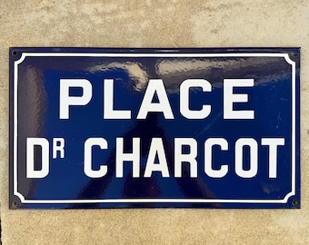 May include: A rectangular blue street sign with white text that reads "PLACE DR CHARCOT". The sign has a white border and is mounted on a light-colored surface. The sign is likely made of metal.
