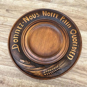 Könnte beinhalten: Eine Holzservierplatte mit einem geschnitzten Design aus Weizen und einer Blume. Die Platte hat einen erhöhten Rand und die Worte "Donnez-Nous Notre Pain Quotidien" sind um den Rand herum geschnitzt.