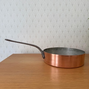 Pode incluir: Uma panela de cobre com uma longa alça curva de metal escuro. A panela tem um exterior de cobre brilhante e um interior prateado. A alça é presa à lateral da panela. A panela está sobre uma superfície de madeira.