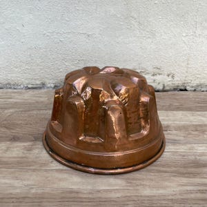 May include: A copper-colored, antique pudding mold with a fluted design. The mold is made of metal and has a rounded shape.