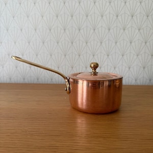 May include: A small copper saucepan with a lid and a long brass handle. The pot has a rounded body and a small brass knob on the lid. The handle is attached to the side of the pot. The pot is sitting on a wooden surface.