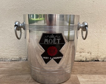 May include: A silver champagne bucket with two ring handles. The bucket features the Moët & Chandon logo on the front, with a black and red emblem. The bucket is made of a shiny, reflective metal and is designed to hold a bottle of champagne.