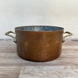 May include: A vintage copper pot with brass handles. The pot has a worn, aged patina and is suitable for cooking or decorative purposes.