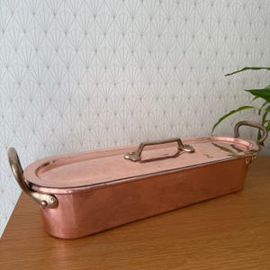 Puede incluir: Una cacerola rectangular de cobre con tapa y asas de latón. El plato tiene un acabado pulido y está diseñado para cocinar. Las asas son curvas y están unidas a los lados, y la tapa tiene un solo asa en el centro.