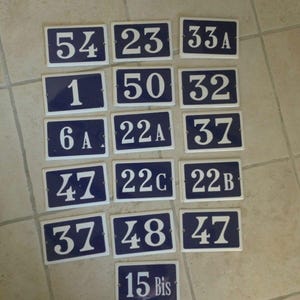 May include: A collection of rectangular blue and white house number signs. The numbers and letters are white on a blue background. The numbers include 1, 6A, 15 Bis, 22A, 22B, 22C, 23, 32, 33A, 37, 47, 48, and 50.