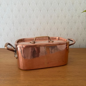 Op de afbeelding: Een rechthoekige koperen kookpot met een deksel en twee zijhandgrepen. De pot heeft een gehamerde textuur en een messing handvat op het deksel. De pot staat op een houten oppervlak.