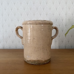 May include: A cylindrical ceramic pot with a beige, glazed finish and two side handles. The vessel features a thick, rounded rim and a slightly textured surface with visible imperfections, showing a darker, unglazed base at the bottom.