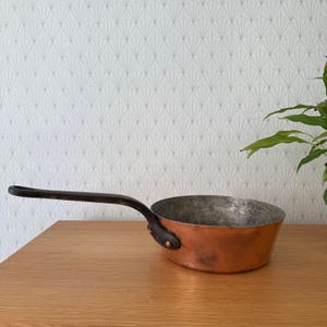 May include: A copper saucepan with a long, dark handle. The pan has a rounded shape and a shiny, metallic interior. The handle is attached with rivets. The pan sits on a wooden surface.