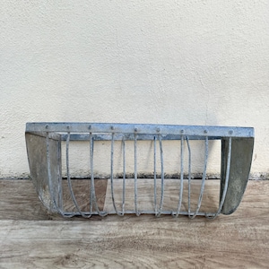 Puede incluir: Un macetero de metal galvanizado con forma curva y diseño de malla de alambre. El macetero está vacío y listo para ser llenado con plantas.