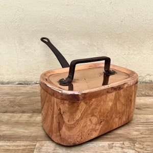May include: A vintage copper pot with a black iron handle. The pot has a rounded rectangular shape and a smooth, shiny surface.