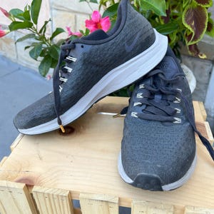 K&ouml;nnte beinhalten: Ein Paar schwarze Nike Laufschuhe mit wei&szlig;en Sohlen. Die Schuhe haben ein Obermaterial aus strukturiertem Stoff und ein schwarzes Nike Swoosh Logo an der Seite.