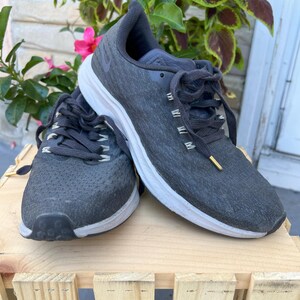 K&ouml;nnte beinhalten: Ein Paar schwarze Nike Laufschuhe mit wei&szlig;en Sohlen. Die Schuhe haben ein strukturiertes Obermaterial und einen schwarzen Schn&uuml;rverschluss.