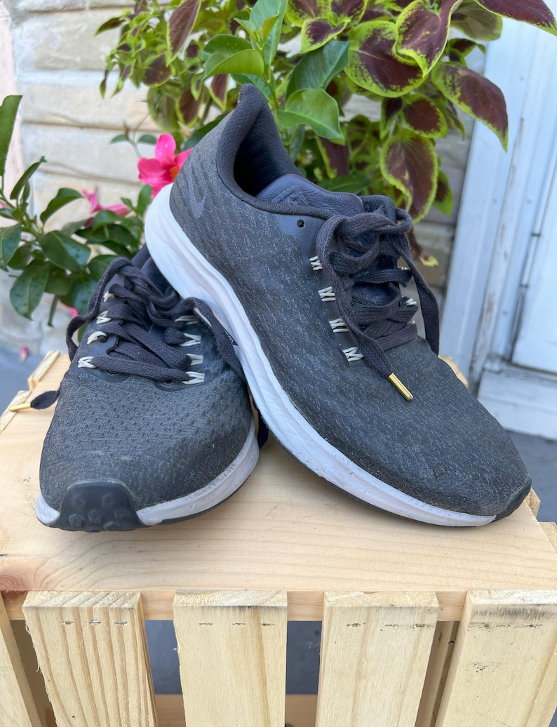 K&ouml;nnte beinhalten: Ein Paar schwarze Nike Laufschuhe mit wei&szlig;en Sohlen. Die Schuhe haben ein strukturiertes Obermaterial und einen schwarzen Schn&uuml;rverschluss.