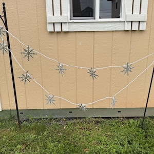 May include: A string of white LED lights featuring silver snowflake-shaped covers, suspended between two black metal shepherd hooks. The decorative light strand displays multiple snowflake ornaments evenly spaced along the white wire cord.