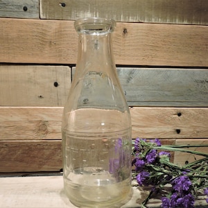 May include: A clear, vintage glass milk bottle with a narrow neck and a wide base. The bottle has etched markings and is set against a rustic wooden background. A sprig of purple flowers rests near the bottle.