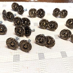 May include: A collection of bronze-colored cabinet knobs with a floral design. Each knob has a central circular element surrounded by detailed, curved petal-like shapes. The knobs are arranged on a light-colored, textured surface.