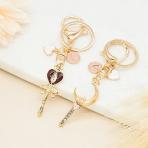 May include: Two gold keychains with charms. One charm is a wand with a pink heart and a pearl, the other is a crescent moon with a pearl. Both charms have rhinestones. Each keychain has a small heart charm and a round charm with a letter.