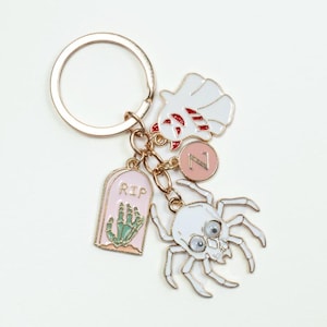 May include: A metal keychain featuring a gold-tone ring attached to four charms: a white ghost with red accents, a pink circular charm with the letter Z, a pink tombstone charm with the text RIP and a green skeletal hand, and a white spider with googly eyes.