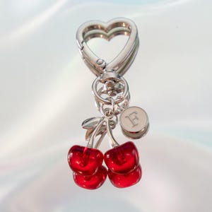 May include: A silver-toned keychain featuring a heart-shaped clasp, a circular charm engraved with the letter F, and two translucent red cherry pendants with silver stems attached to a central ring, reflecting on a smooth surface.