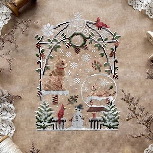 May include: A cross-stitch pattern on tan fabric titled Four Seasons Gazebo Series Winter Gazebo by Stitch with Coffee Designs. The design features a gazebo, a cat, a snowman, cardinals, and snowflakes in red, green, white, and brown thread.