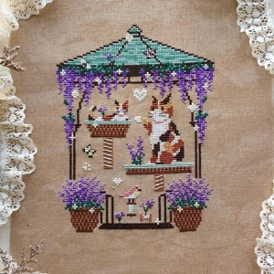 May include: A cross-stitch pattern titled Summer Gazebo from the Four Seasons Gazebo Series, featuring a calico cat and a smaller cat inside a gazebo adorned with purple wisteria, potted lavender, and a small bird, stitched on tan fabric.