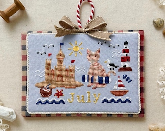 May include: A cross-stitch design for July 2025 featuring a sandcastle, a lighthouse, a cat, and a seagull. The design is on a light blue background with the word "July" stitched in yellow. The calendar is trimmed with a red and white checkered pattern.