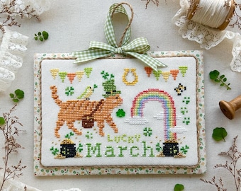 May include: A cross-stitch pattern of a cat wearing a green hat and holding a pot of gold. The cat is standing in front of a rainbow and a horseshoe. The text "Lucky March" is stitched below the cat. The pattern is framed and hanging from a green and white gingham ribbon.