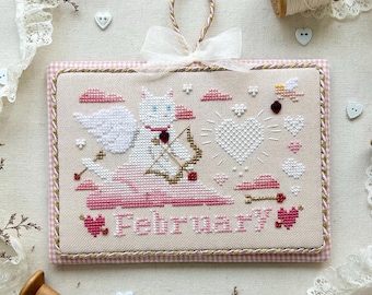 May include: A cross-stitch pattern featuring a white cat dressed as Cupid with a bow and arrow, flying through the air with pink clouds and hearts. The pattern is titled "February" and is part of a monthly kitty calendar series for 2025.