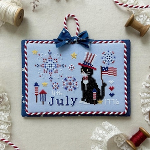 May include: Cross-stitch design for July 2025, featuring a black cat in a patriotic hat holding an American flag. The design includes fireworks and the text "July 1776". The frame is blue with red and white accents.