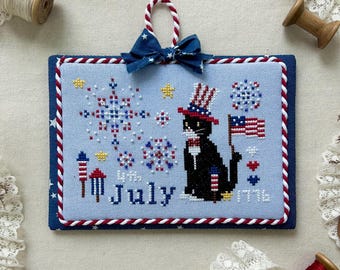 May include: Cross-stitch design for July 2025, featuring a black cat in a patriotic hat holding an American flag. The design includes fireworks and the text "July 1776". The frame is blue with red and white accents.
