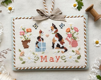 May include: A cross-stitch pattern featuring a cat with a watering can and flowers. The pattern is framed in a light blue and white frame with a brown and white border. The text "May" is stitched in the center of the pattern. The text "Stitch with Coffee Designs" and "May 2025" are printed below the pattern.