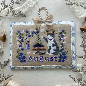 以下が含まれることがあります： 帽子をかぶった白黒の猫が、青とピンクの花の刺繍に囲まれたクロスステッチのアートワーク。猫の下には「August」の文字が刺繍されています。アートワークは青い花のボーダーとベージュの背景で額装されています。「August 2025」の文字が下に表示されています。