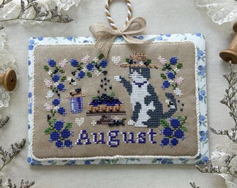 May include: Cross-stitch artwork featuring a black and white cat wearing a hat, surrounded by blue and pink floral embroidery. The word "August" is stitched below the cat. The artwork is framed with a blue floral border and a beige background. The text "August 2025" is displayed below.