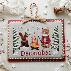 Könnte beinhalten: Ein Kreuzstichdesign mit einer Winterszene mit einem Eichhörnchen und einem Fuchs, die Marshmallows an einem Lagerfeuer rösten. Das Wort "Dezember" ist rot gestickt. Das Design ist mit einem roten Rand und einer beigen Schleife umrahmt. Der Text "Dezember 2025" befindet sich unten.