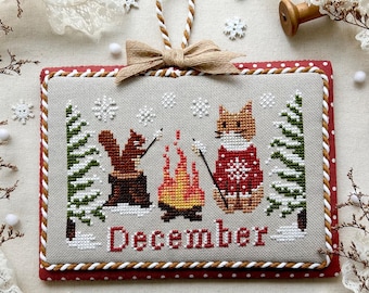 May include: A cross-stitch design featuring a winter scene with a squirrel and fox roasting marshmallows by a campfire. The word "December" is stitched in red. The design is framed with a red border and a beige bow. The text "December 2025" is at the bottom.