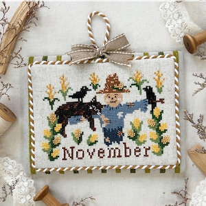 May include: A cross-stitch calendar for November 2025, featuring a scarecrow, crows, and corn. The design includes the text "November" and "Monthly Kitty Calendar Series 2025". The calendar is framed with a green and white border and has a decorative bow.