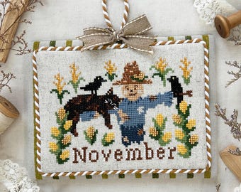 May include: A cross-stitch calendar for November 2025, featuring a scarecrow, crows, and corn. The design includes the text "November" and "Monthly Kitty Calendar Series 2025". The calendar is framed with a green and white border and has a decorative bow.