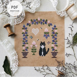 May include: Cross stitch pattern featuring a black cat sitting under a blueberry arch with pink flowers and bees. The pattern is on a beige fabric and includes the text "Tuxey's Blueberry Arch" and "Stitch with Coffee Designs".