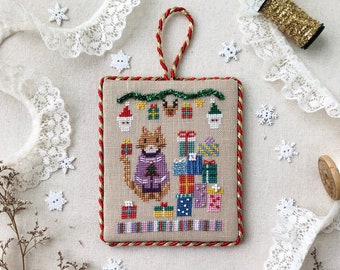 May include: Cross stitch ornament featuring a cat wearing a Christmas sweater and surrounded by presents. The ornament is framed with a red and gold trim. The text "Stitch with Coffee Designs" and "Christmas Sweater Series Christmas Gift-Giving" is below the ornament.