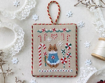 May include: A cross-stitched Christmas ornament featuring a bunny wearing a blue sweater, surrounded by candy canes and snowflakes. The ornament is framed with red and gold thread, with a matching hanging loop. Text reads "Christmas Sweater Series Candy Canes."