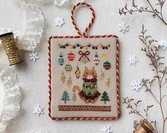 May include: A cross-stitch ornament featuring a cat wearing a Christmas sweater with a Christmas tree design. The cat is surrounded by ornaments and a string of lights. The ornament is framed with a red and gold border. The text "Stitch with Coffee Designs" and "Christmas Sweater Series Christmas Ornaments" is below the ornament.