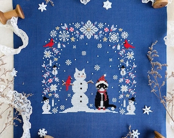 May include: Cross stitch pattern featuring a black cat and a snowman with a red cardinal bird. The design is surrounded by a wreath of snowflakes, holly, and other winter elements. The text "Kitty, It's Cold Outside" is displayed below the image. Stitch with Coffee Designs is the name of the designer.