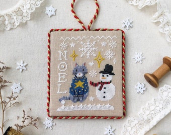 May include: A rectangular, framed cross-stitch ornament featuring a cat and snowman design. The cat wears a blue sweater with a star, and the snowman has a black hat and red scarf. The word "NOEL" is stitched above the figures, with snowflakes and stars.