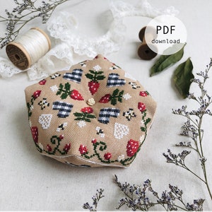 Puede incluir: Un biscornu beige de punto de cruz con un patrón de fresas y cuadros vichy. El biscornu está rodeado de encaje blanco, carretes de hilo y ramitas de flores secas. El texto "Strawberry Biscornu" y "Stitch with Coffee Designs" está impreso debajo del biscornu. El texto "www.stitchwithcoffee.etsy.com" está impreso debajo del nombre del diseño.