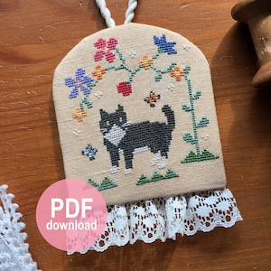 Könnte beinhalten: Ein Kreuzstichmuster eines schwarzen Katers mit weißen Abzeichen, der in einem Garten mit Blumen und Schmetterlingen sitzt. Das Muster ist auf einem beigefarbenen Stoff mit einer weißen Spitzenborte. Das Muster ist als PDF-Download erhältlich.