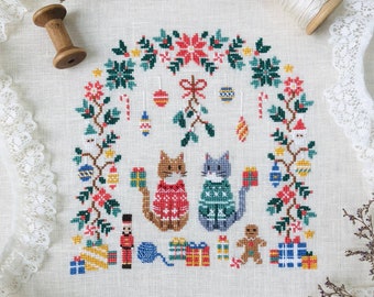 May include: Cross stitch pattern featuring two cats wearing sweaters under a mistletoe archway with Christmas decorations. The pattern includes a gingerbread man, a nutcracker, and presents. The text "Stitch with Coffee Designs" and "Under the Mistletoe" is included in the image.