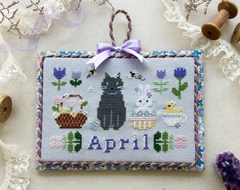 May include: Cross stitch pattern featuring a black cat, a white bunny, and a yellow chick. The animals are surrounded by flowers and a basket of eggs. The design is titled "April" and is part of a monthly kitty calendar series.