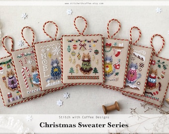May include: A set of six cross-stitch ornaments featuring cats wearing Christmas sweaters. Each ornament is a square with a red and white border and a beige background. The ornaments feature different designs, including a cat wearing a sweater with ornaments, a cat wearing a sweater with candy canes, a cat wearing a sweater with the word "NOEL", a cat wearing a sweater with a reindeer, a cat wearing a sweater with a snowman, and a cat wearing a sweater with the word "JOY".