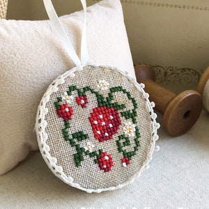 May include: A round, cream-colored ornament with a white lace trim. The ornament is embroidered with a red and green strawberry design. The ornament has a white ribbon attached for hanging.