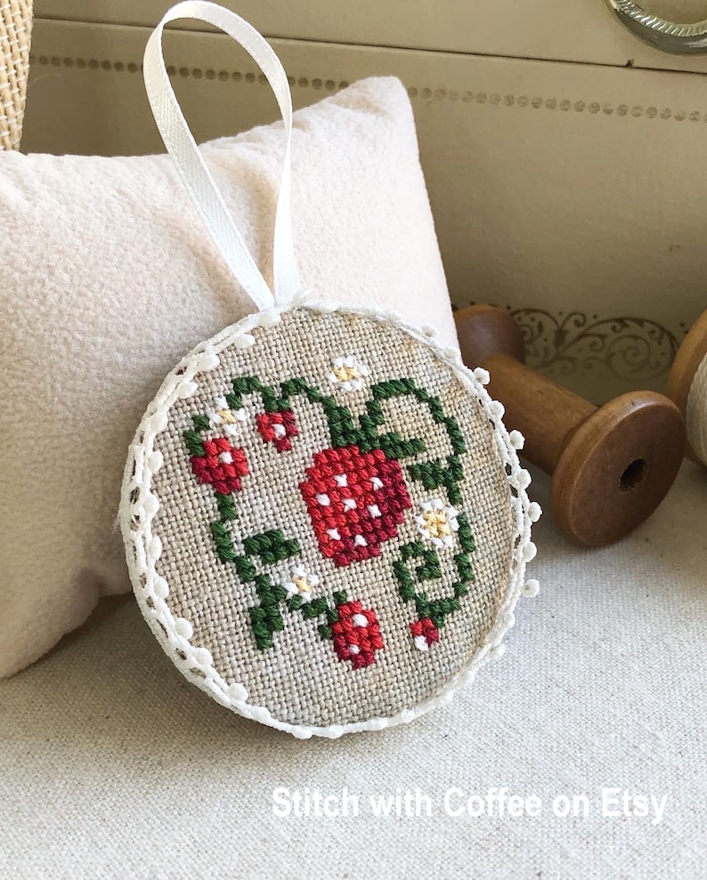 May include: A round, cream-colored ornament with a white lace trim. The ornament is embroidered with a red and green strawberry design. The ornament has a white ribbon attached for hanging.